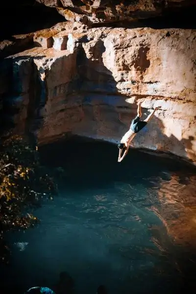 A shirtless man mid-dive from a warm sandstone cliff into a green-blue pool at an Agadir Morocco surf cove