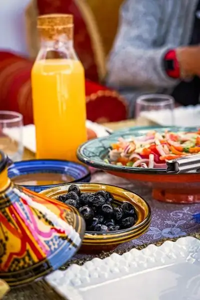A low, patterned table set with Moroccan dishes including a painted tagine lid, glossy black olives, a bright tomato salad, and a carafe of orange juice on the Agadir Morocco Surf coastline.