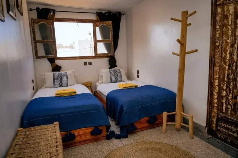 Two wooden beds with deep-blue throws and yellow towels in a crisp white room overlooking sunlit rooftops of a beachside town – agadir morocco surf
