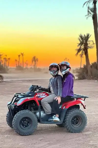 Two riders in helmets on a red KYMCO MXU quad bike at sunset on sandy dunes with palm trees in Anza – agadir morocco surf