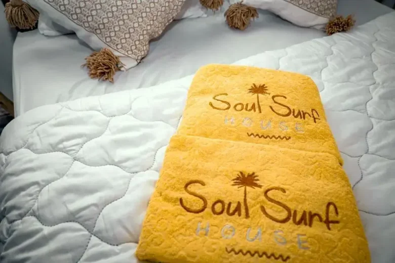 Neatly made bed with crisp white quilt, tasseled pillows, and golden-yellow ‘Soul Surf House’ towels with palm logo - agadir morocco surf