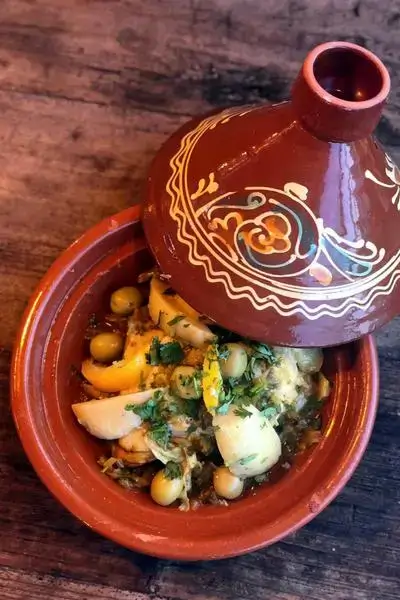 Glazed terracotta tagine pot half-open on a weathered wooden table reveals a colorful chicken stew with green olives, preserved lemon wedges, onions and cilantro under warm light, evoking agadir morocco surf vibes