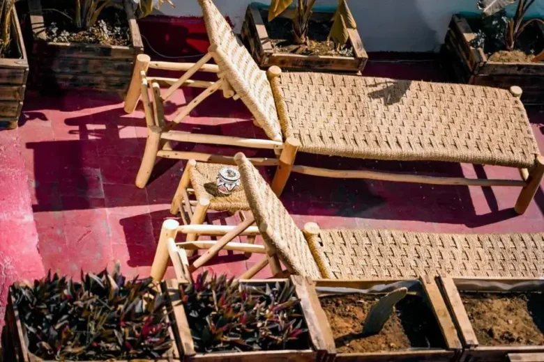 A pair of handcrafted wooden lounge chairs with woven straw seats and backs on a sun-warmed red-tiled terrace, accompanied by a small side stool with a hand-painted ceramic pot and surrounded by rustic planter boxes, agadir morocco surf