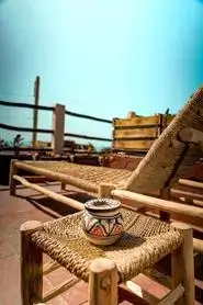 Small handwoven wooden stool with painted ceramic jar, driftwood chaise lounge and weathered crates on sunbaked terracotta tiles under a vivid blue sky in Anza – agadir morocco surf