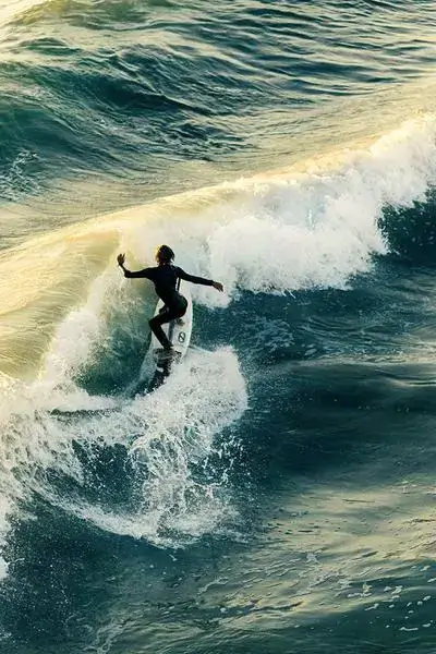 Lone surfer in a full wetsuit carving a rolling wave at Anza beach, golden light glinting off the green-blue ocean, agadir morocco surf