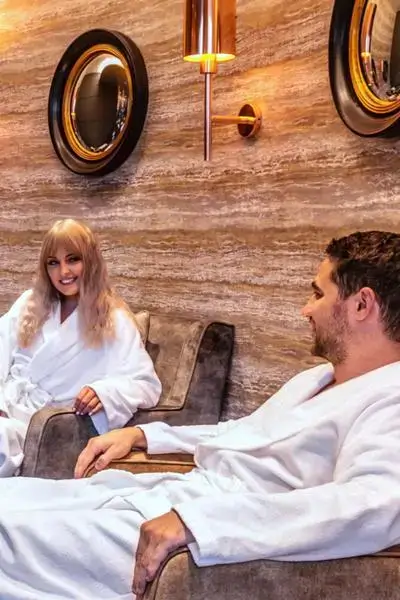 Young couple in white spa robes relaxing in a plush suede lounge with stone walls, copper sconces, and circular mirrors after surfing in Anza agadir morocco surf