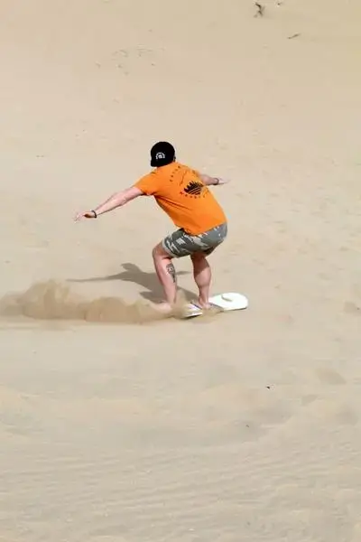 A barefoot rider in an orange T-shirt and camo shorts leans on a sandboard down a sunlit dune, sand spraying behind him in Anza, Morocco — agadir morocco surf
