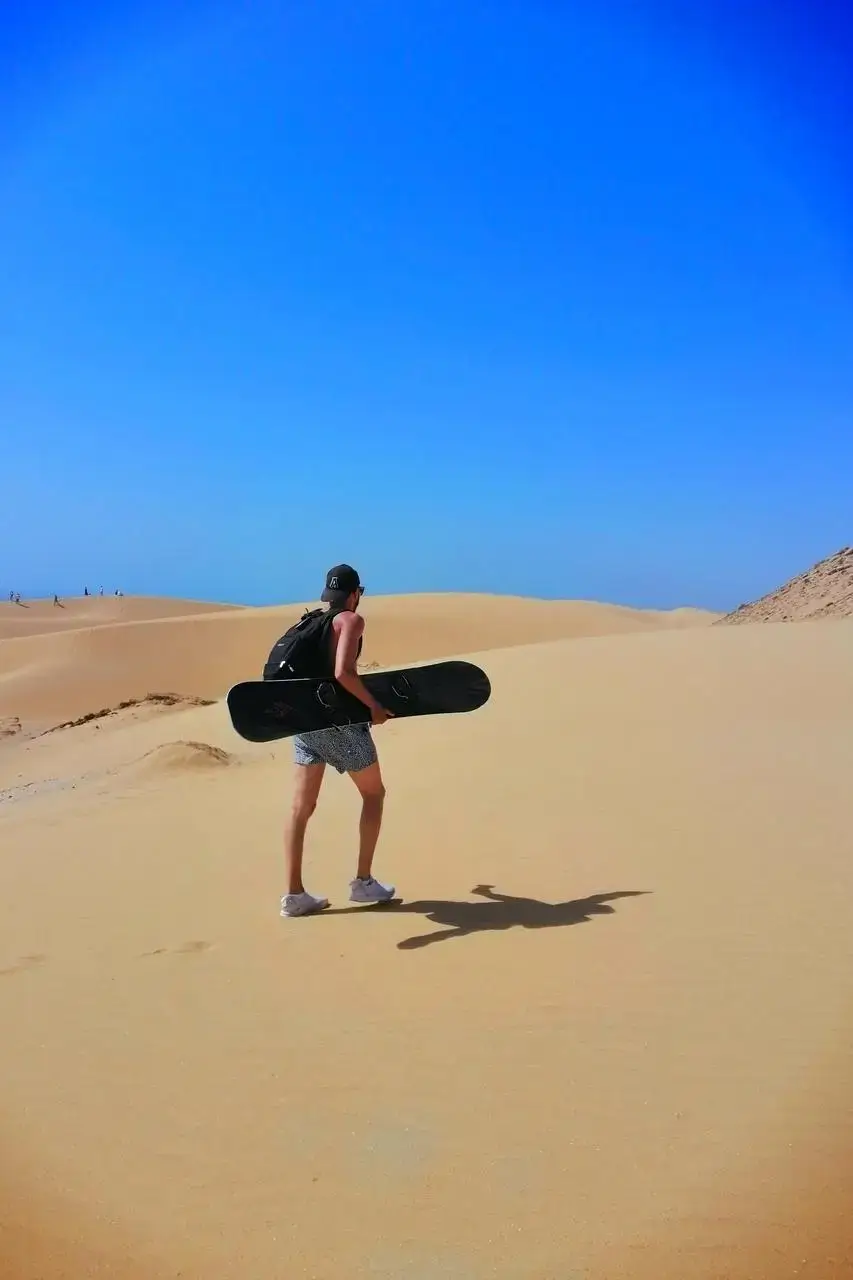Lone adventurer with a black sandboard climbing golden dunes under intense blue sky, agadir morocco surf