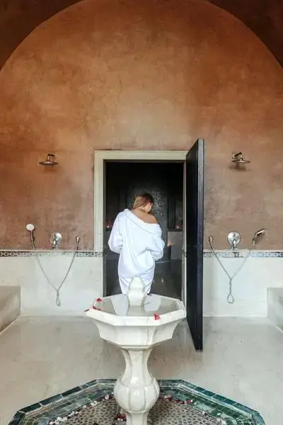 Lone figure in a plush white robe stepping through a dark-framed arch into a Moroccan hammam spa chamber with central marble fountain and rose petals, agadir morocco surf