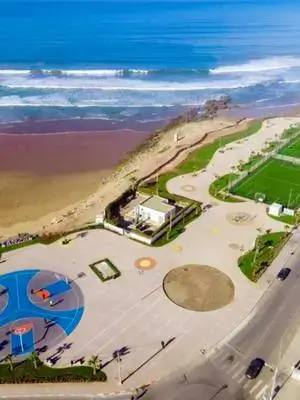 Aerial view of Anza seaside promenade with a blue basketball court, soccer pitch, palm-lined walkways, and surfers riding Atlantic waves. agadir morocco surf