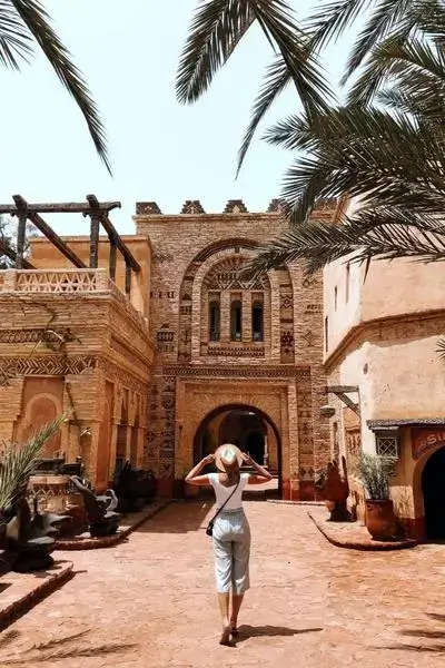 A lone visitor in light summer attire and a straw hat walking down a terracotta-paved lane toward an ornately carved mudbrick gateway framed by palm fronds in agadir morocco surf