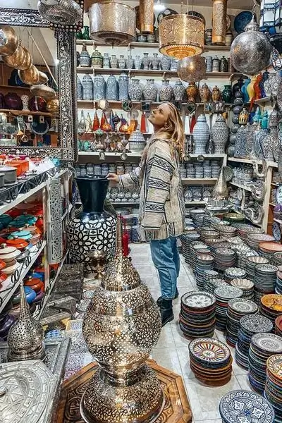 A young woman in denim and a surf-style jacket browses hand-painted tagine bowls and ceramics under hanging lanterns in a coastal Moroccan shop – agadir morocco surf