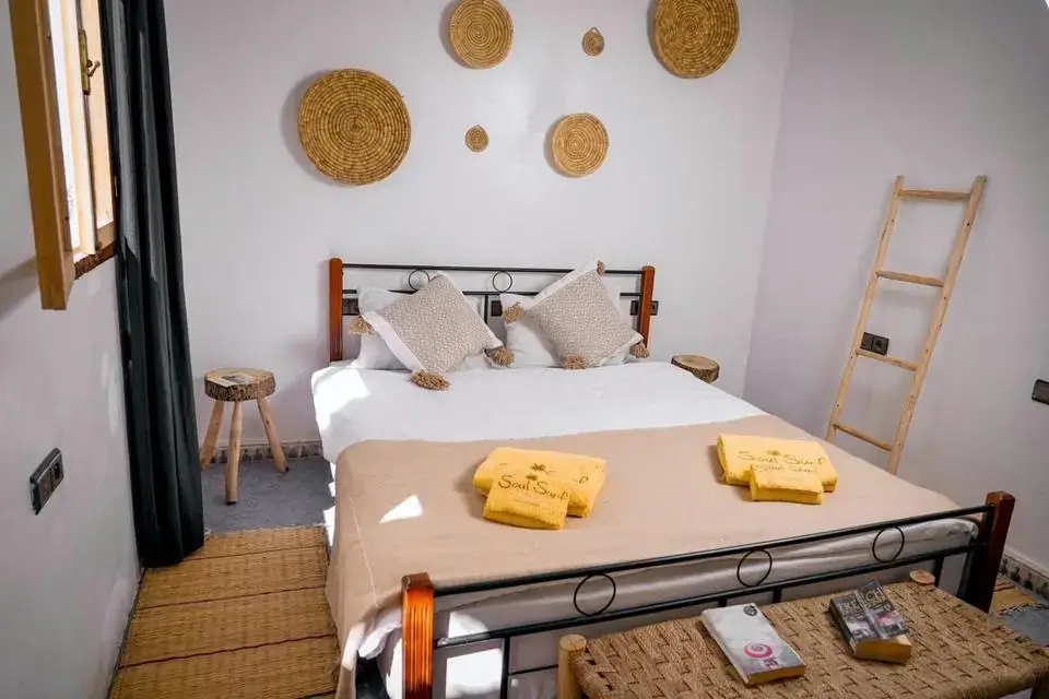 agadir morocco surf cozy bedroom with metal-frame bed, crisp white linens, sandy-beige blanket, handwoven straw plates, and surf towels