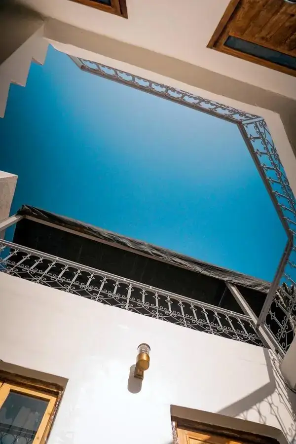 agadir morocco surf – sunlit whitewashed courtyard with polished wooden shutters, ornate wrought-iron balcony, brass sconce, and geometric metal trellis against a cerulean sky