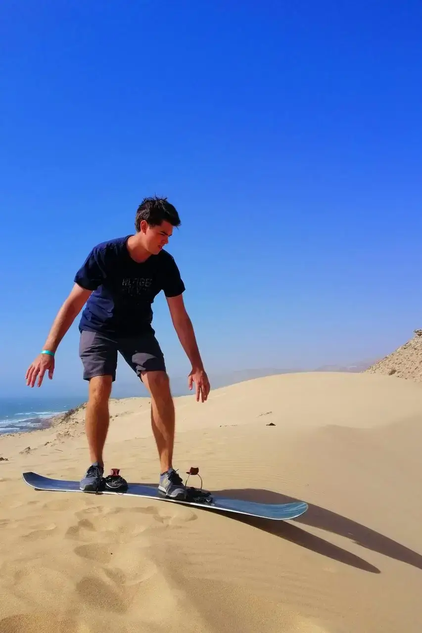 Young man stands poised on a sandboard at the crest of an Anza dune with pale Atlantic waves below agadir morocco surf