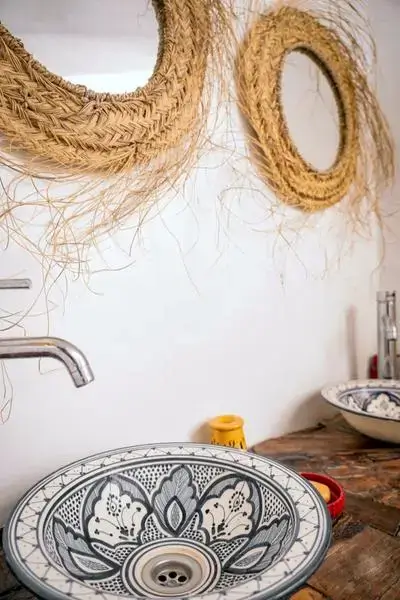 Hand-painted Moroccan ceramic basin on weathered wooden counter with straw-framed round mirrors, mustard-yellow candle holder, and red dish – agadir morocco surf