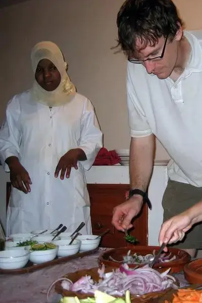 Young woman in hijab and chef’s jacket and man in polo preparing fresh herbs and vegetables on a table indoors under warm light at Anza beach, agadir morocco surf setting